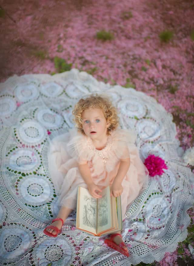 Featured on Cake and Lace pretty in pink Park tea party small girl wearing pink dress looking up to the camera on white blanket with pink petals on the ground