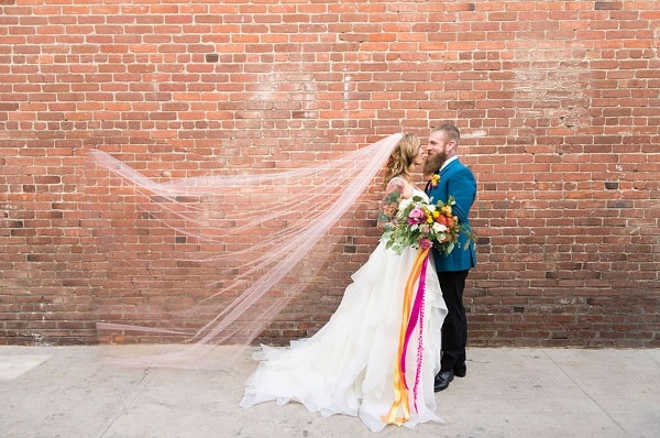 bride and groom looking at each other in front of a brick wall with veil floating in the wind