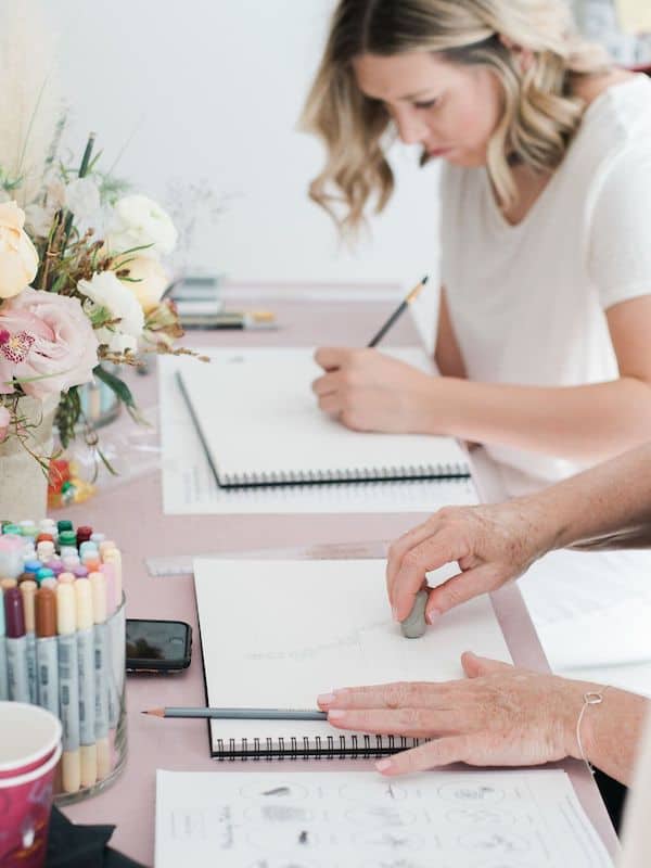 hads with an eraser on paper and an individual drawing in a sketchbook in the background