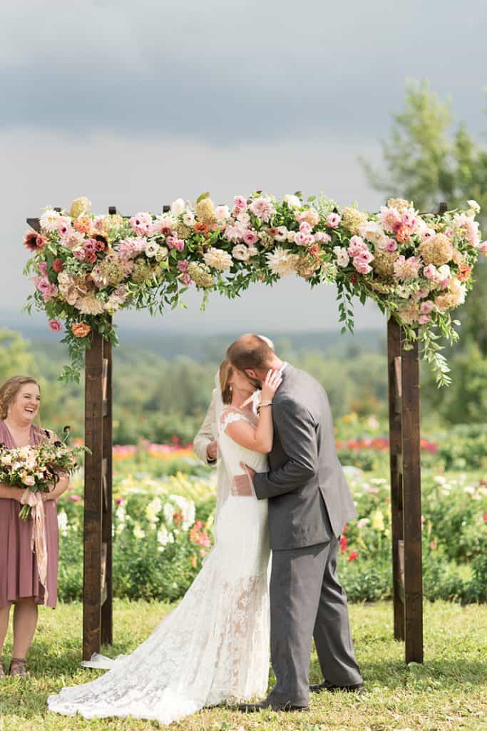 bride and groom kissing under wedding arch decorated with peach pink and white flowers