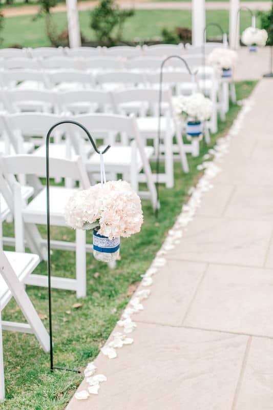 Aisle lined with white flower petals, handing hydrangeas and white chairs on grass
