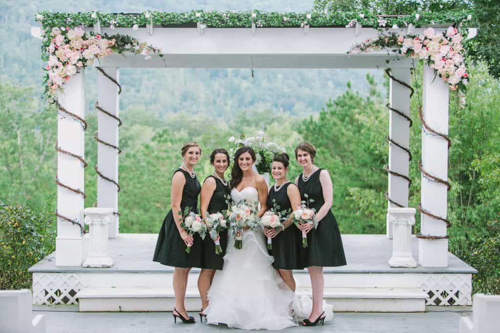 bride wearing white and bridsmaids in black standing in front of large white gazebo decorated with pink and white florals
