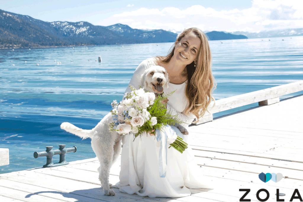 bride holding flowers and dog on a dock with water and mountain background