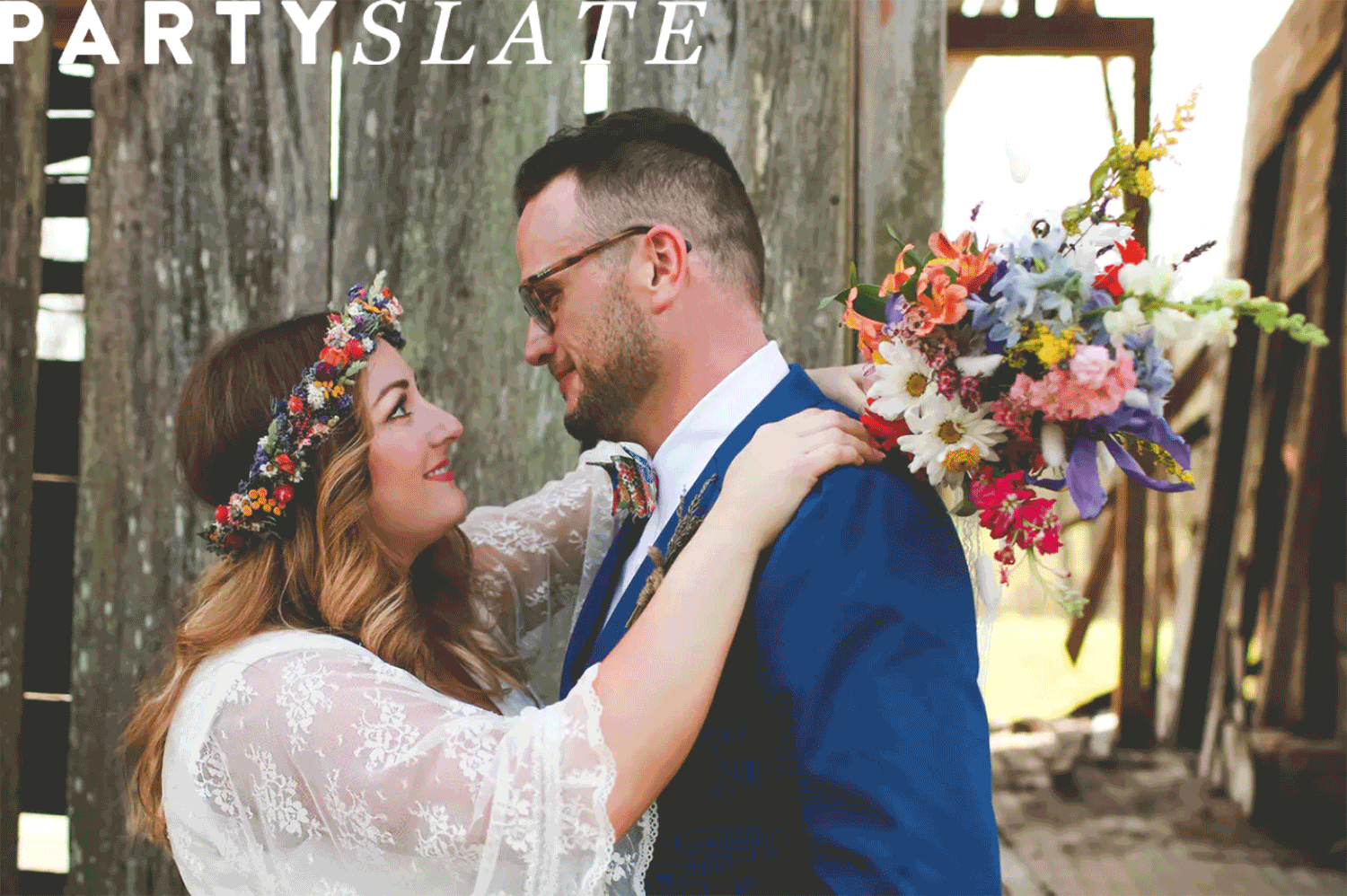 bride with multi-colored floral crown gazing at husband outdoors in front of wooden fence boards