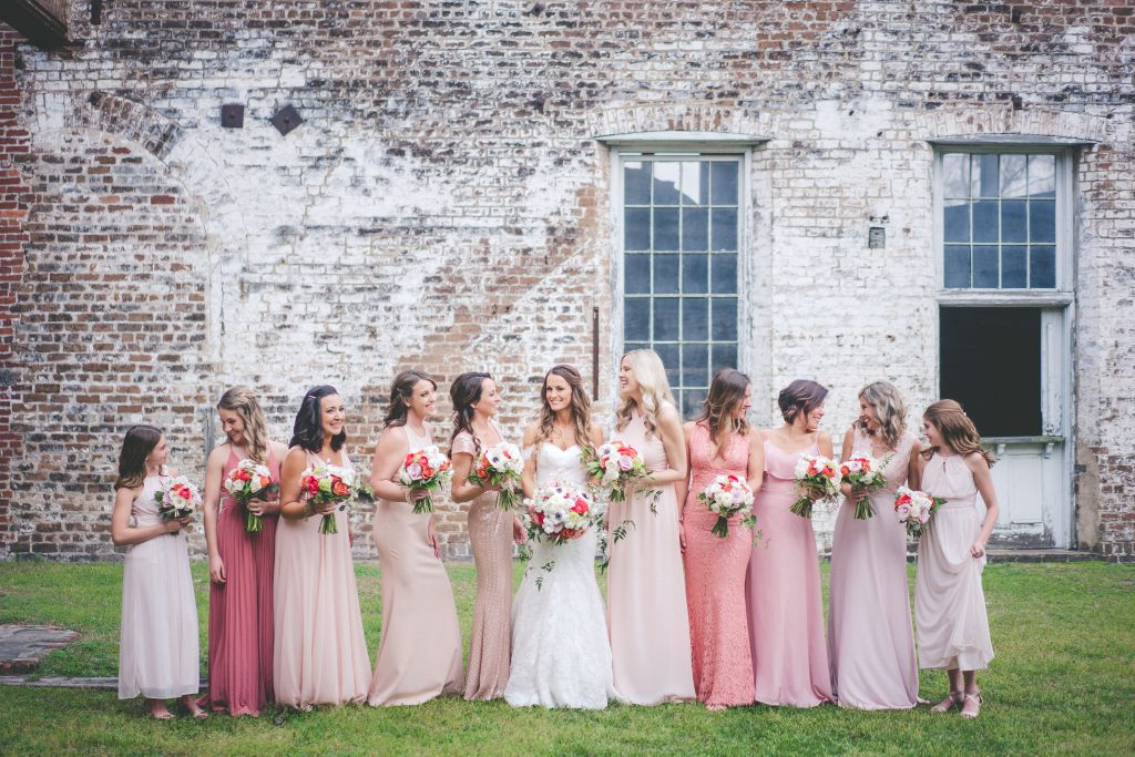 Bride and bridesmaids wearing pink, purple and peach dresses holding flowers in front of brick wall