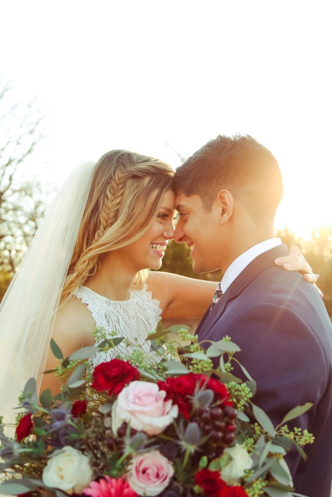 Smiling bride and groom outdoors with the sun int he background and flowers in the foreground