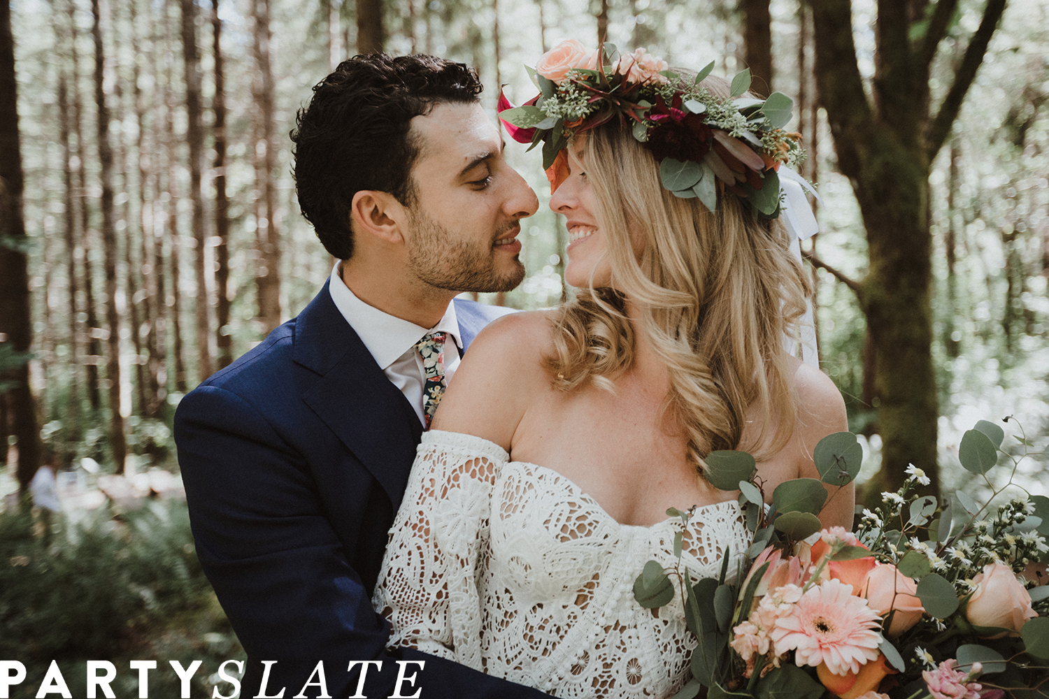 Bride wearing floral crown and holding bouquet of flowers in the forest facing groom