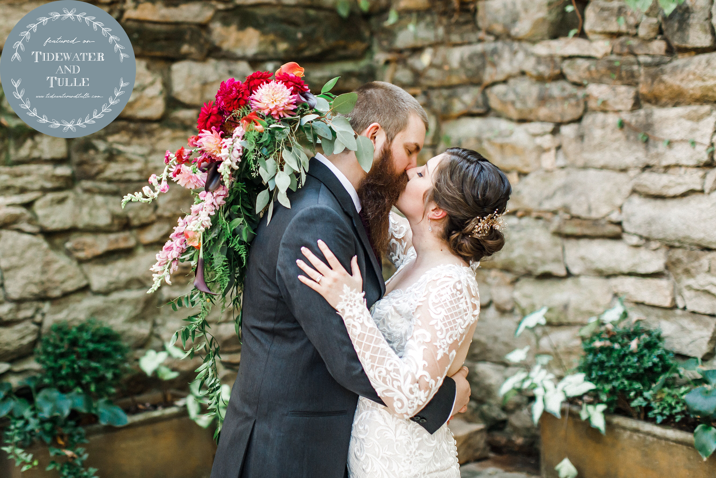 Wedding couple kissing with pink and red floral bouquet and stone wall in the background