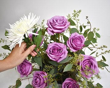 hand adding white football mums to purple flowers with seeded eucalyptus stems in floral foam