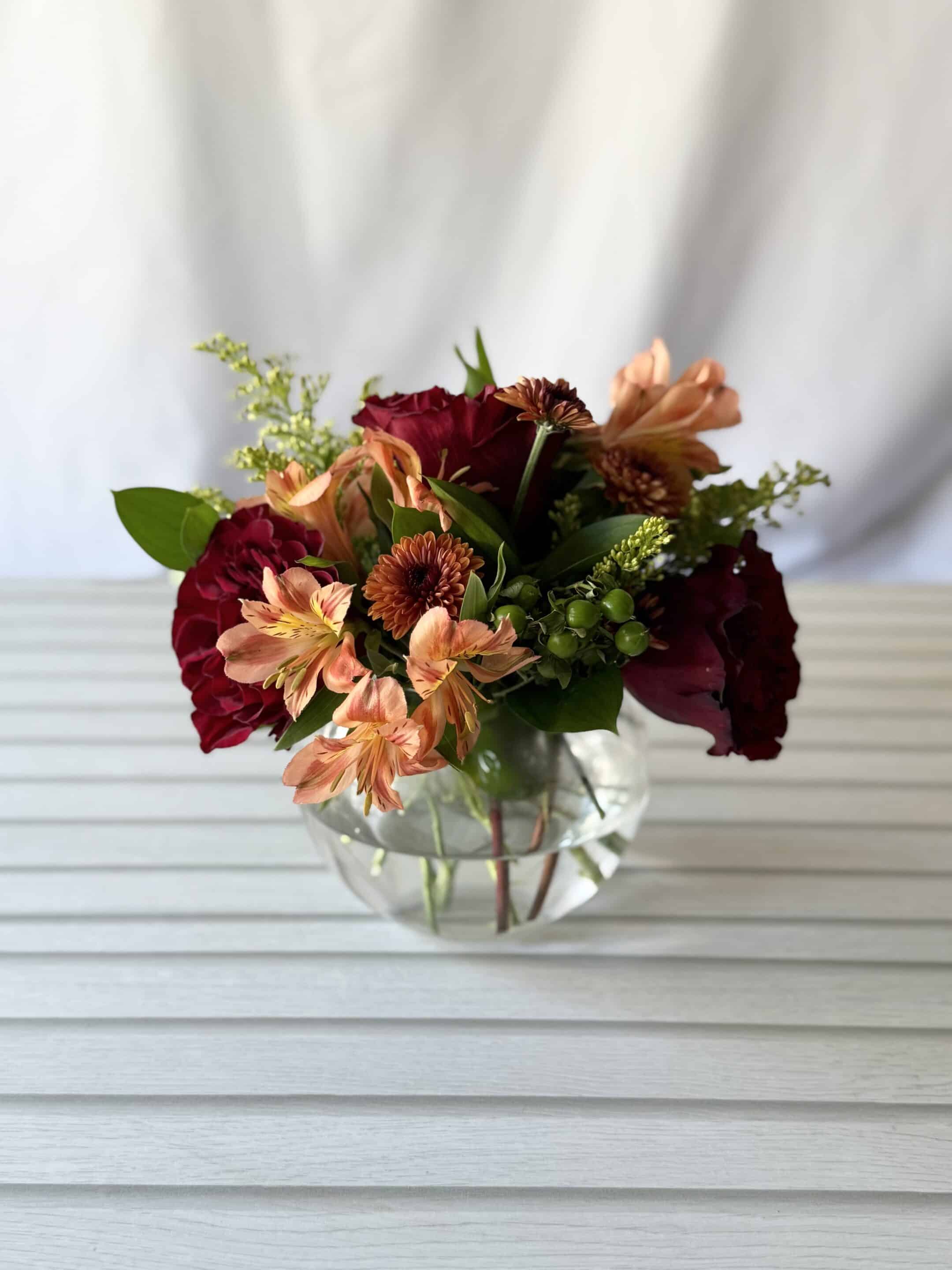 orange alstromeria, greenery and red roses in a small round glass vase