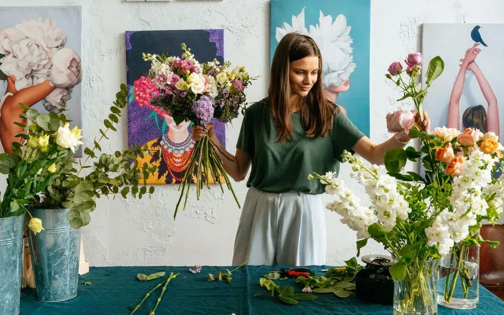 Woman holding a mixed bouquet and arranging roses beside buckets of fresh flowers.