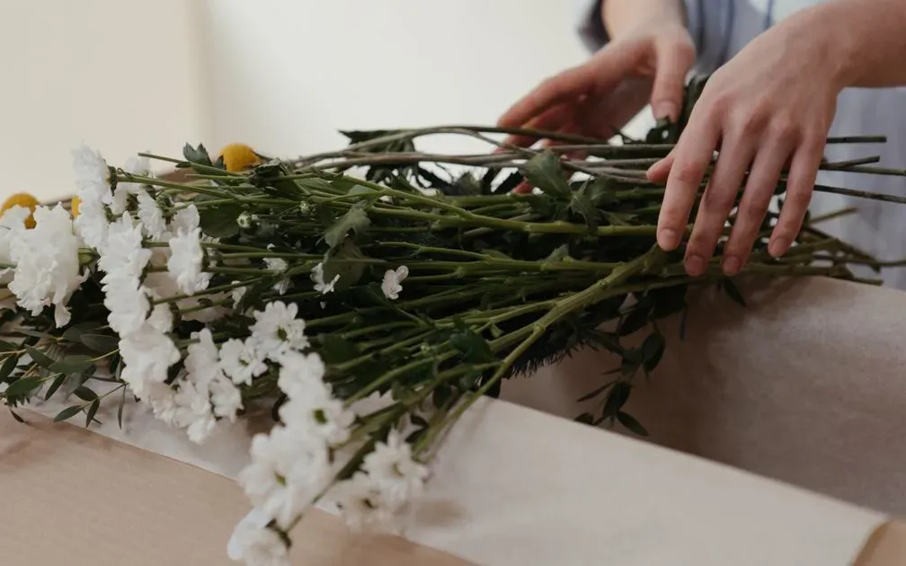 Hands arranging a bundle of white flowers and greenery on brown kraft paper.