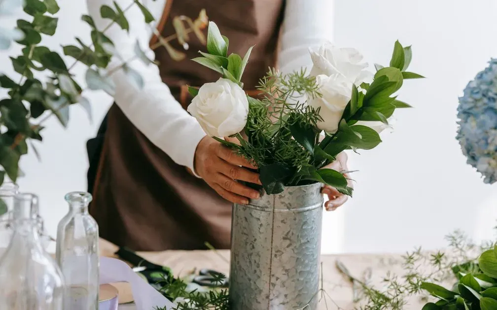 A florist in an apron arranges white roses and greenery in a metal bucket.