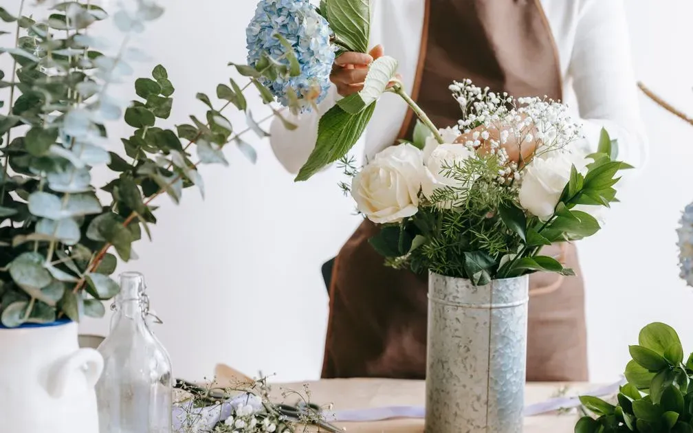 Florist in a brown apron adds blue hydrangea to a vase of white roses and greenery.