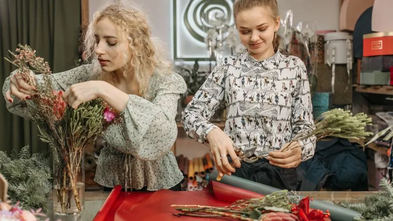 Two women arranging dried flowers and greenery at a worktable in a flower shop.