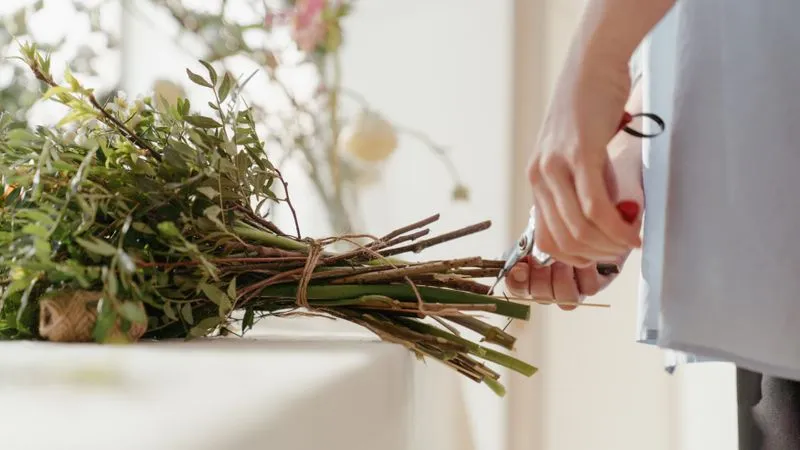 Hands use red-handled pruning shears to trim the stems of a twine-tied greenery bundle.