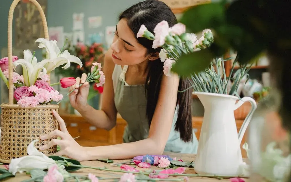Florist in an apron arranging pink carnations, roses and lilies in a wicker basket