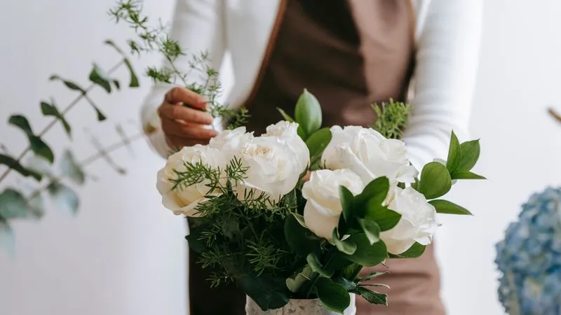 Person in a brown apron arranging white roses and fresh greenery in a vase