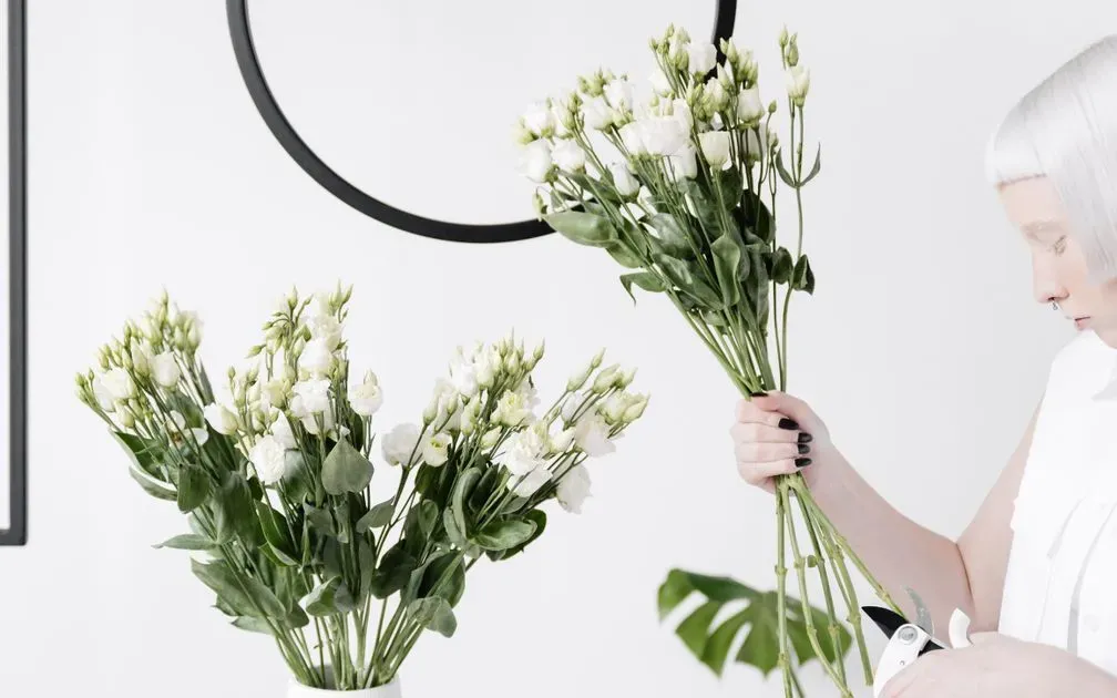 A woman holding a bunch of white lisianthus stems and pruning shears beside a vase.