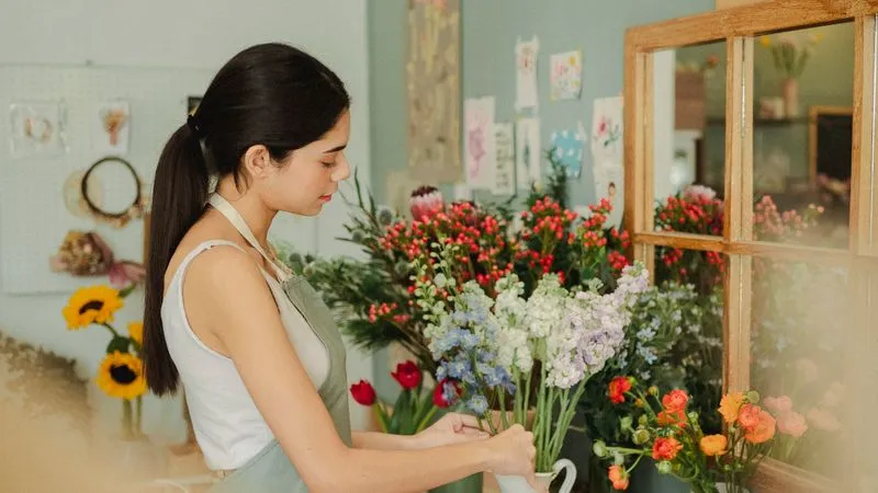 Florist in an apron arranging fresh blue, white and lilac flowers in a white pitcher.