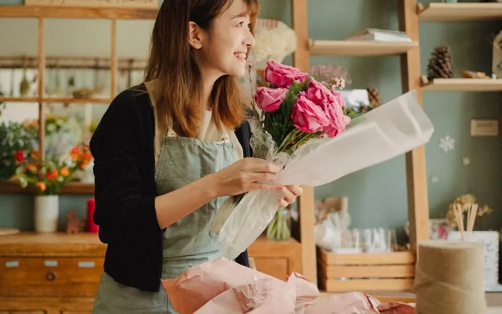 Smiling florist in an apron wrapping a bouquet of pink roses in a flower shop.