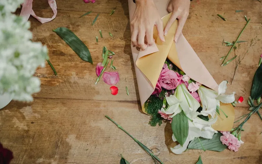 Hands wrapping a bouquet of lilies, roses and carnations in paper on a wooden table.