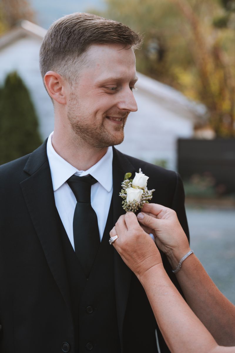 Hands pin a boutonniere of two white spray roses and seeded eucalyptus to a groom's black suit lapel.