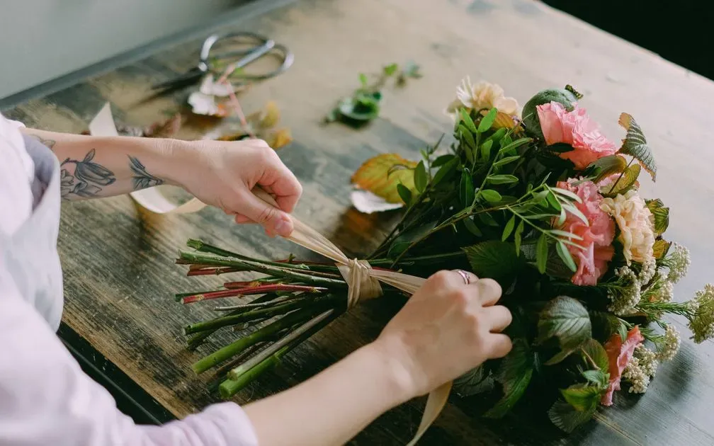 Florist ties a ribbon around a bouquet of pink roses and greenery on a wooden table.