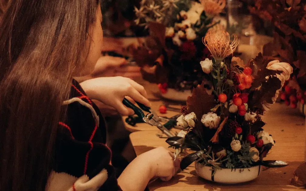 A woman trims stems with shears beside an autumn arrangement of roses, berries and leaves.